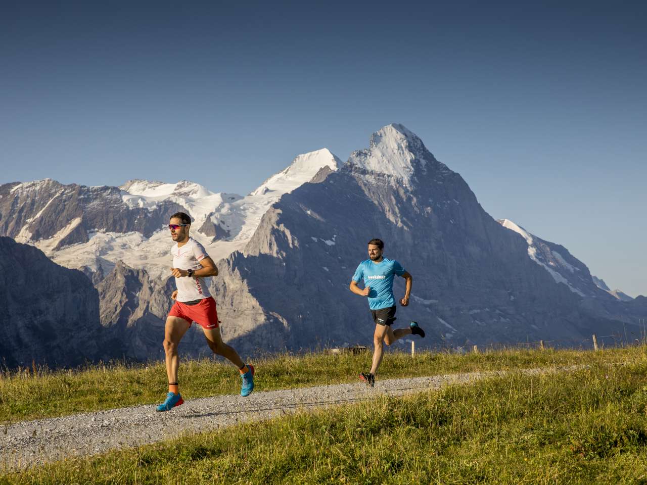 Zwei Menschen beim Trail Running auf einem Bergpfad, umgeben von alpiner Landschaft mit Felsen, Wiesen und Bergen im Hintergrund.
