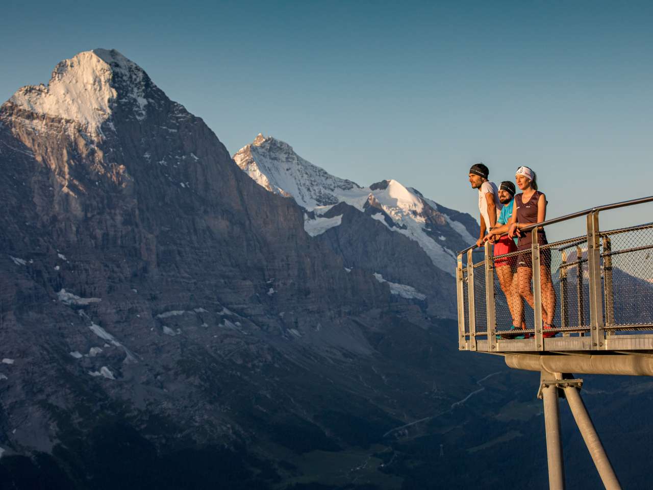 Drei Menschen stehen auf einer Aussichtsplattform, im Hintergrund eine beeindruckende Bergkulisse unter klarem Himmel.