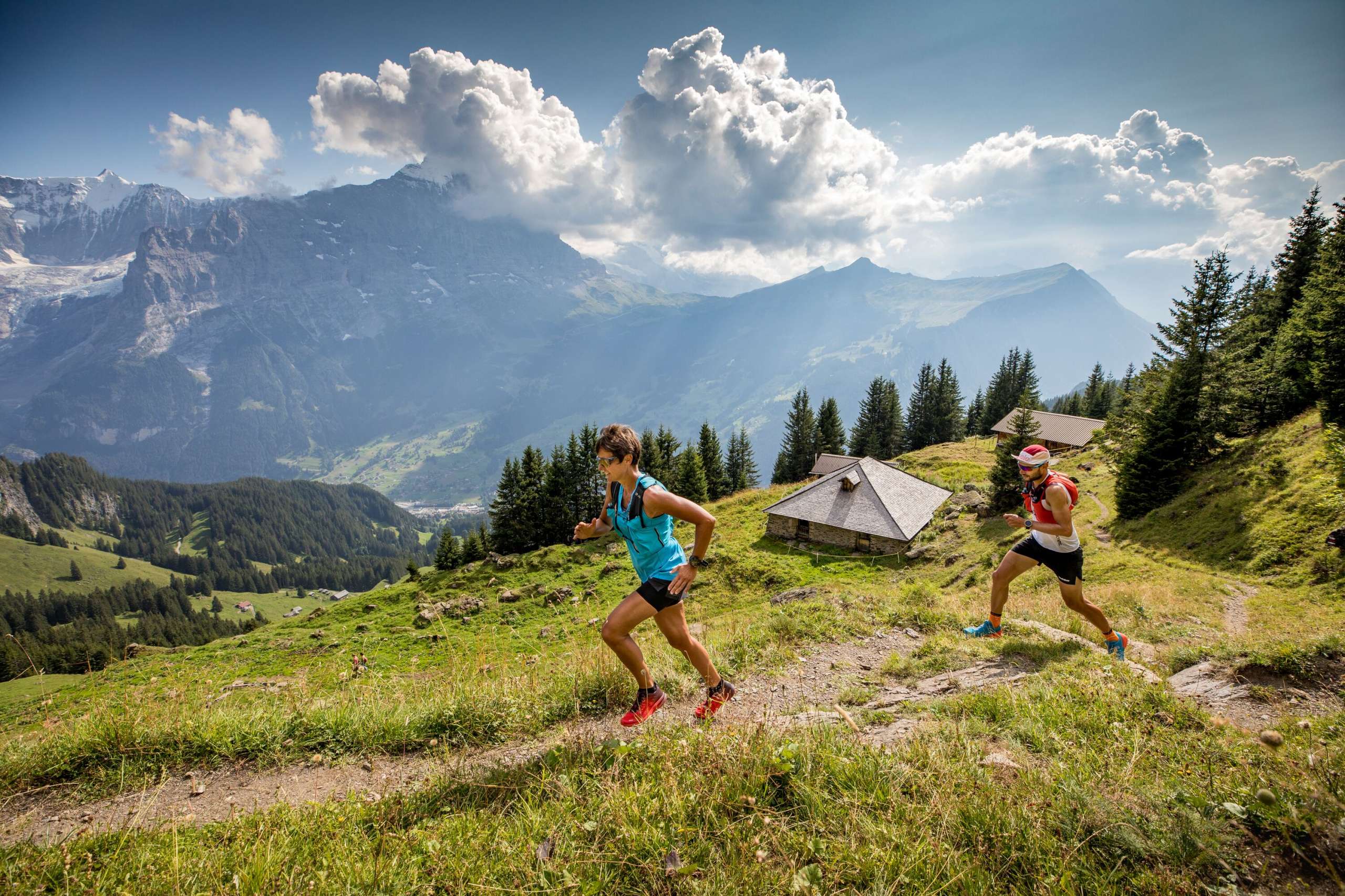 Zwei Menschen beim Trail Running auf einem Bergpfad, umgeben von alpiner Landschaft mit Felsen, Wiesen und Bergen im Hintergrund.